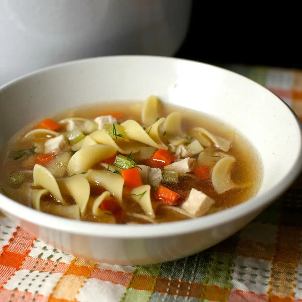 A bowl of chicken noodle soup is on a plaid piece of cloth.