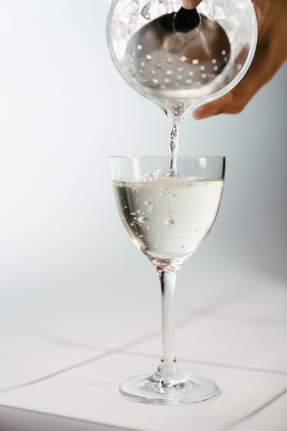 hand pouring clear liquid from a drink shaker into a wine glass