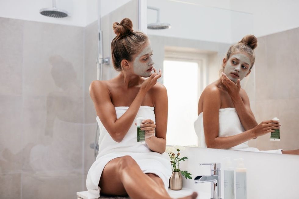 Young woman sitting on the sink counter applying a face mask while looking in a mirror. 